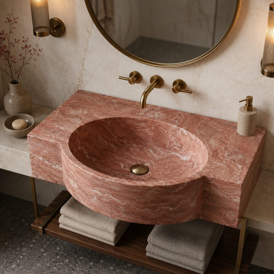 A round vessel sink made of pinkish-red marble with natural veining, mounted on a matching marble countertop with brass fixtures, set in a modern bathroom.