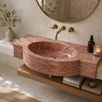 A modern bathroom vanity featuring a carved reddish-brown marble sink and matching countertop, mounted on a wooden floating shelf. The setup includes brass wall-mounted faucets, a round mirror, a lit candle, a soap dispenser, and folded towels on the shelf below.