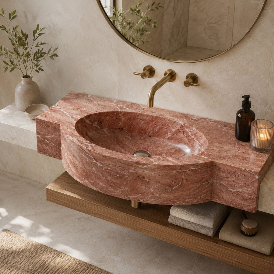 A modern bathroom vanity featuring a carved reddish-brown marble sink and matching countertop, mounted on a wooden floating shelf. The setup includes brass wall-mounted faucets, a round mirror, a lit candle, a soap dispenser, and folded towels on the shelf below.