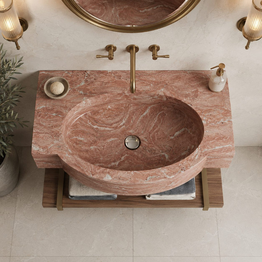 A round pinkish-red marble vessel sink with a matching rectangular countertop, mounted on a wooden floating shelf. The setup includes a brass faucet with two handles, a soap dish with a bar of soap, and a white pump bottle, set against a beige tiled wall with wall sconces and a mirror.