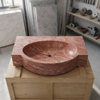 A carved red marble washbasin with a thick rectangular base, displayed on a wooden crate in a workshop setting with marble slabs and tools in the background.