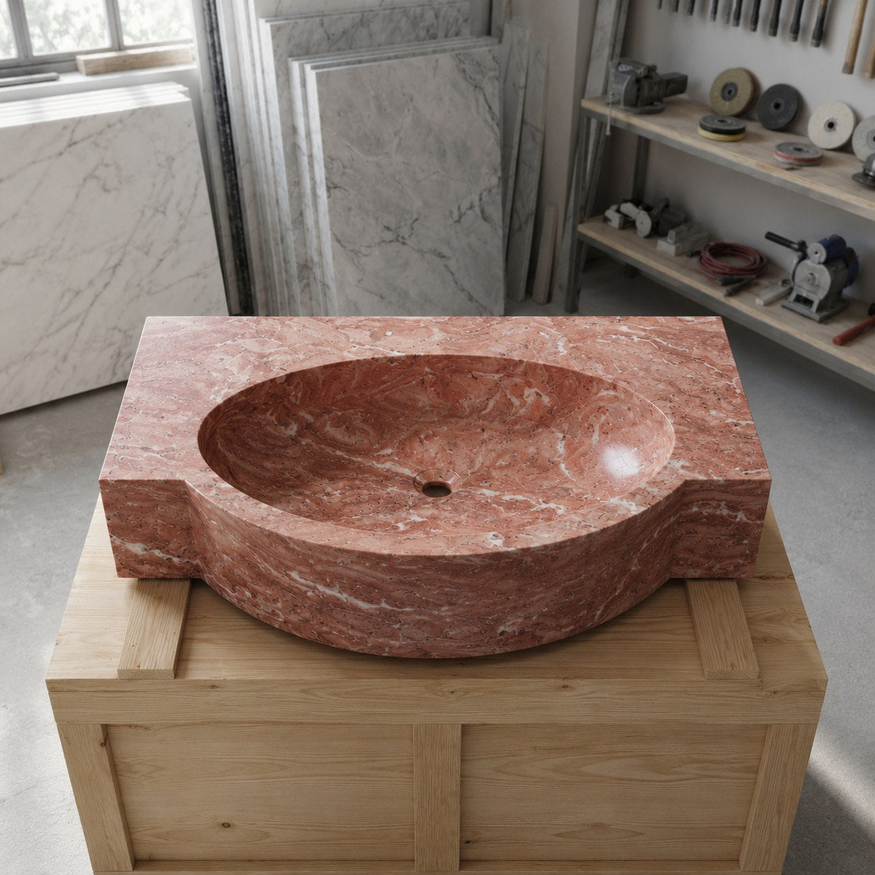A carved red marble washbasin with a thick rectangular base, displayed on a wooden crate in a workshop setting with marble slabs and tools in the background.