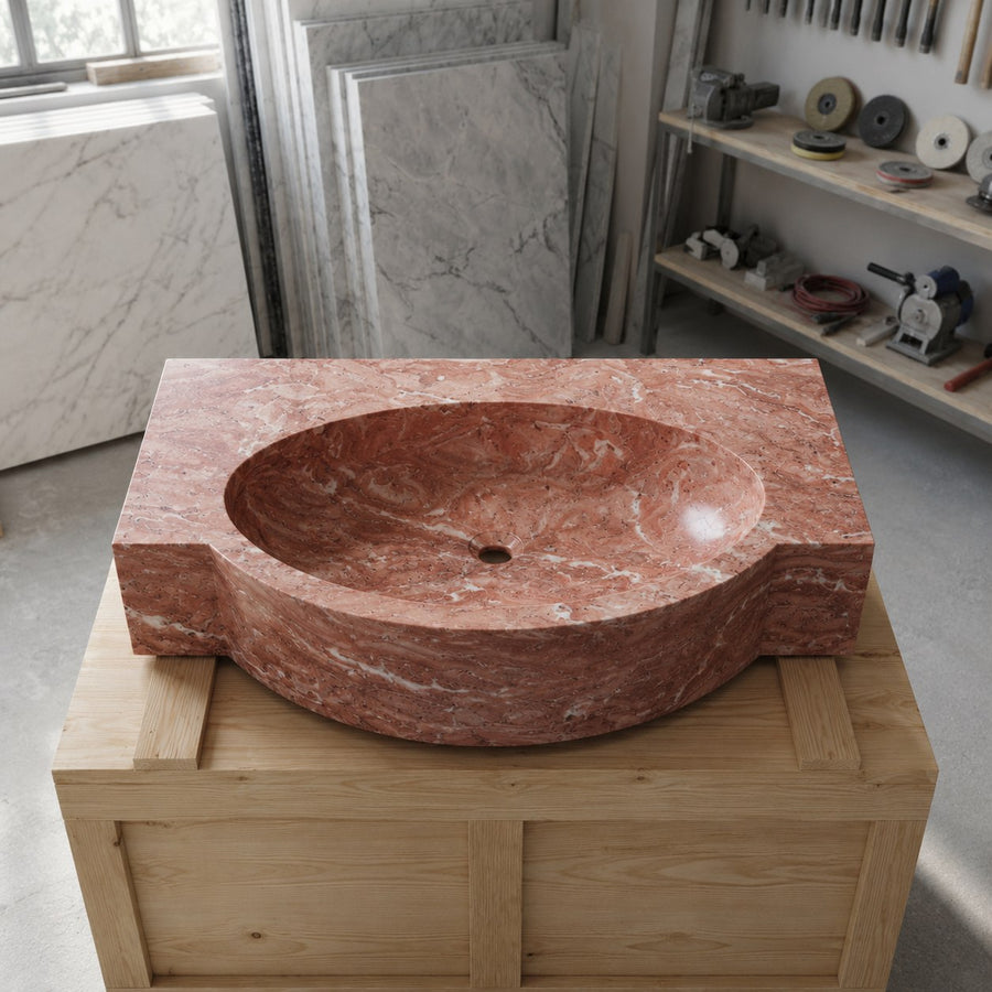 A carved red marble washbasin with a thick rectangular base, displayed on a wooden crate in a workshop setting with marble slabs and tools in the background.