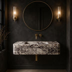A modern floating bathroom sink with a black and white marble pattern, mounted against a dark textured wall. The sink is flanked by two vertical glass sconces and a large round gold-framed mirror above, with gold wall-mounted faucets.