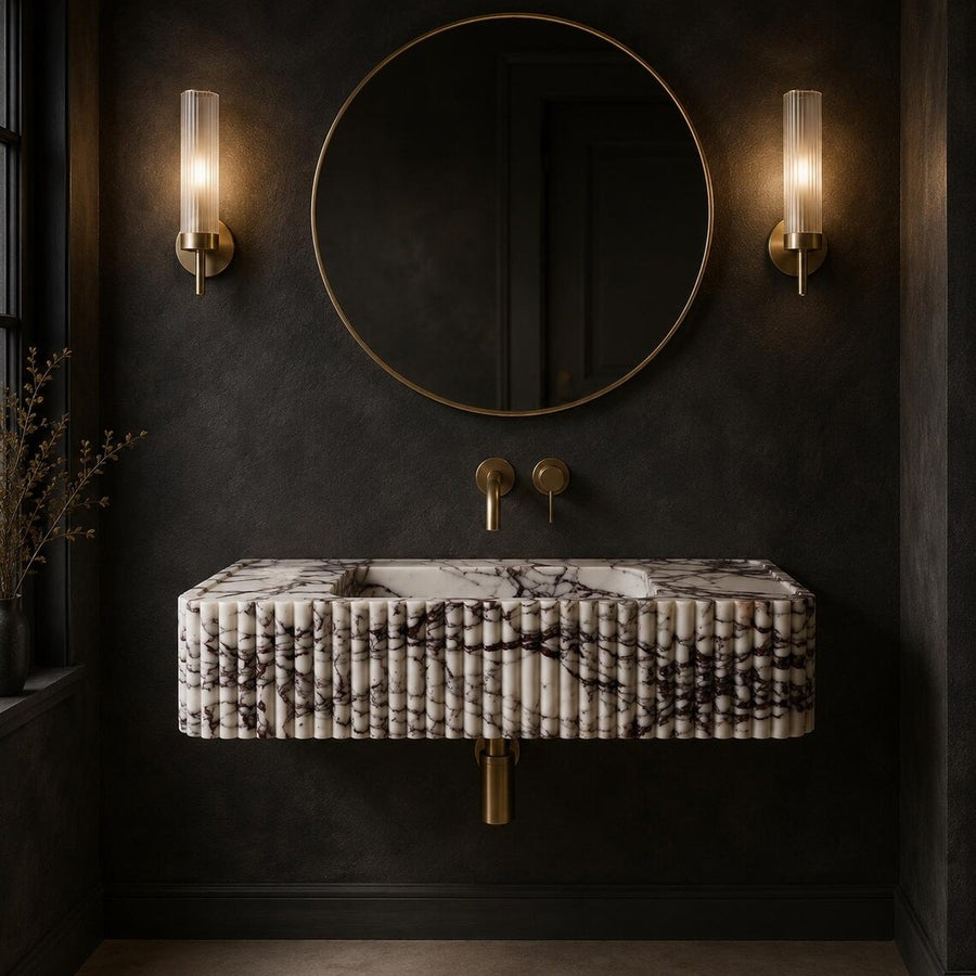 A modern floating bathroom sink with a black and white marble pattern, mounted against a dark textured wall. The sink is flanked by two vertical glass sconces and a large round gold-framed mirror above, with gold wall-mounted faucets.