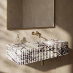 A modern floating bathroom sink with a white marble-look surface and dark veining, mounted against a textured beige wall beneath a matching mirror. A gold faucet and a clear soap dispenser are also visible.