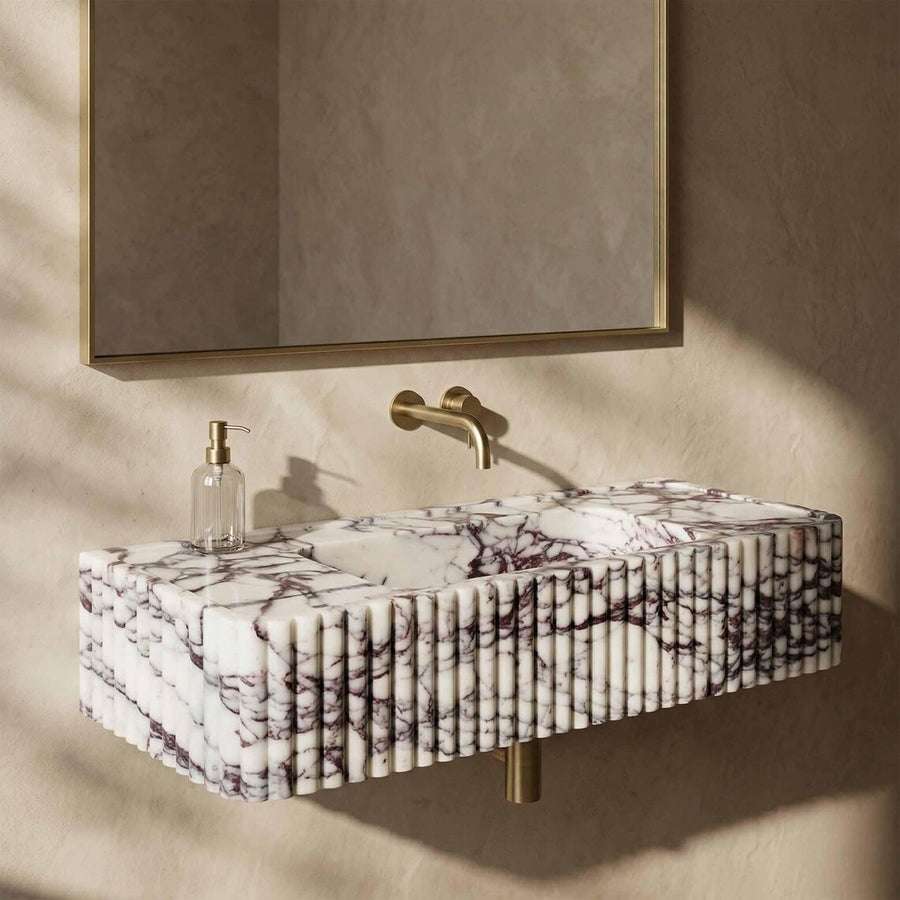 A modern floating bathroom sink with a white marble-look surface and dark veining, mounted against a textured beige wall beneath a matching mirror. A gold faucet and a clear soap dispenser are also visible.
