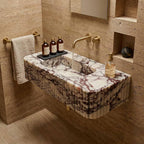 A floating rectangular marble sink with a fluted edge, featuring white and gray veining, mounted on a beige stone wall in a bathroom setting with gold fixtures.