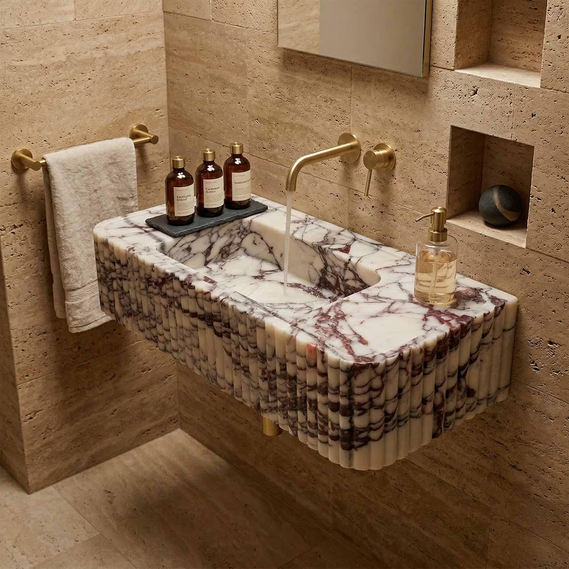 A floating rectangular marble sink with a fluted edge, featuring white and gray veining, mounted on a beige stone wall in a bathroom setting with gold fixtures.