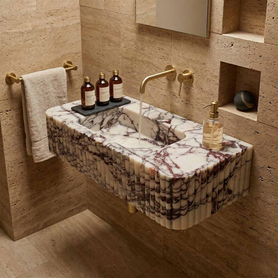A floating rectangular marble sink with a fluted edge, featuring white and gray veining, mounted on a beige stone wall in a bathroom setting with gold fixtures.