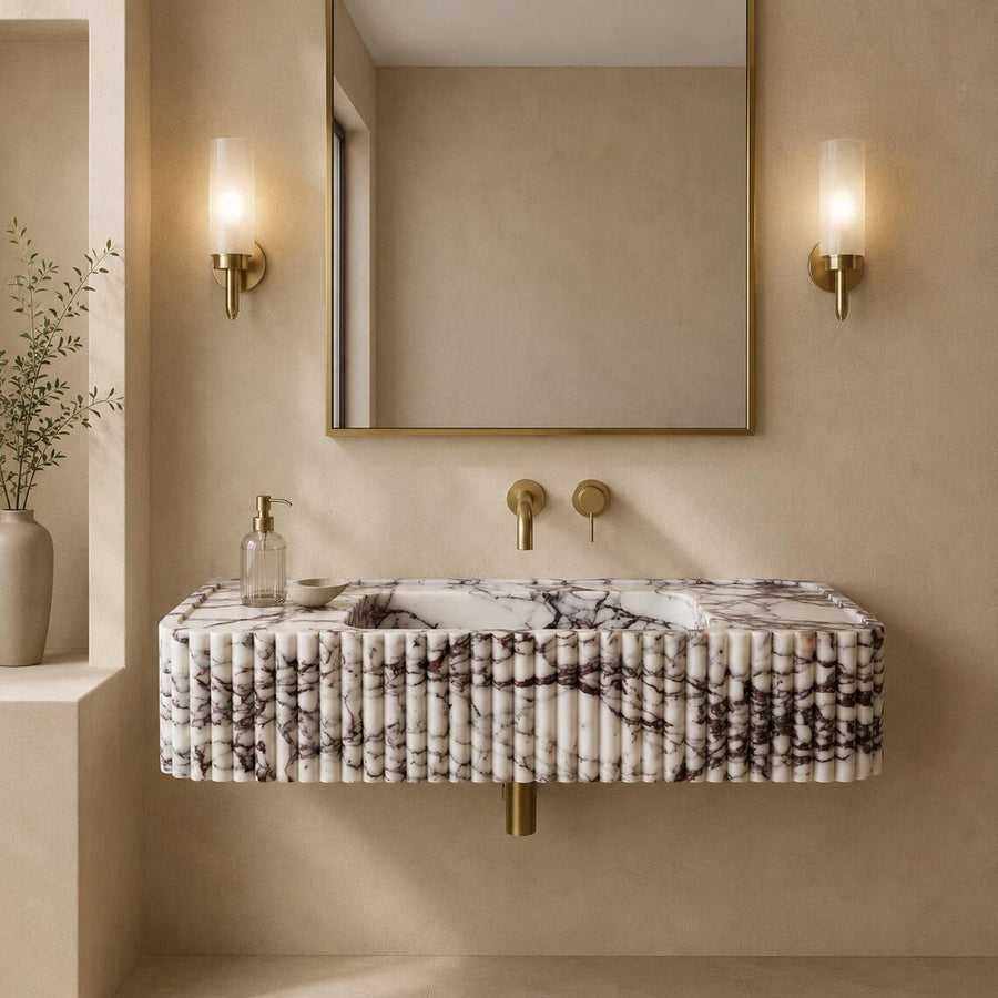 A modern floating vanity with a pleated white marble front and black veining, mounted against a beige plaster wall. A matching rectangular mirror with a gold frame hangs above, flanked by two gold wall sconces.
