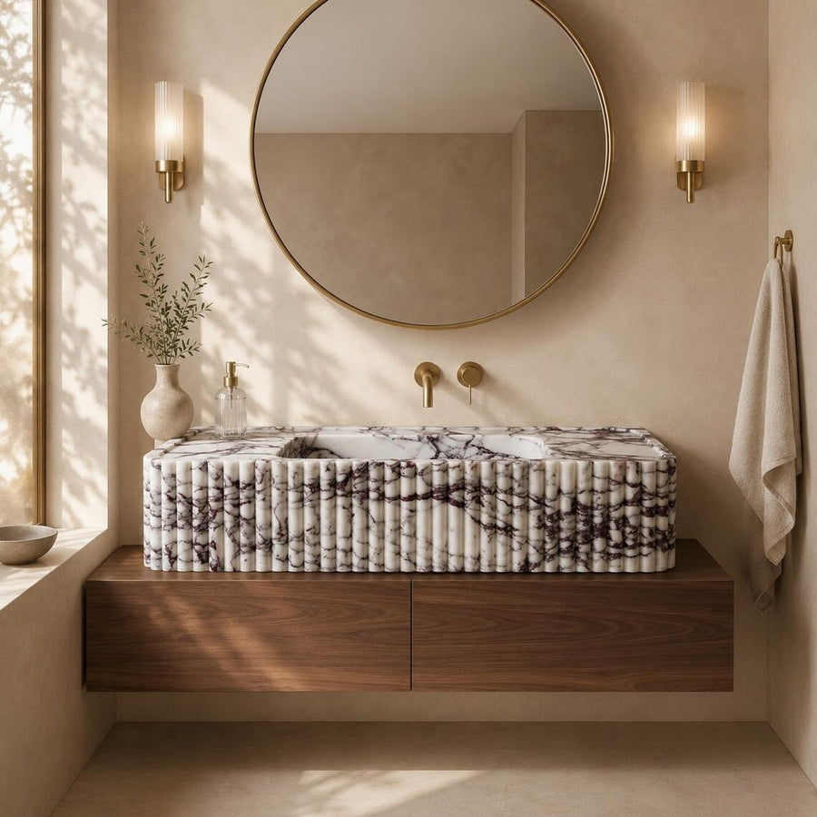 A floating bathroom vanity with a ribbed white marble-look countertop and dark wood cabinet, set in a warm beige bathroom with a round mirror, gold fixtures, and wall sconces.