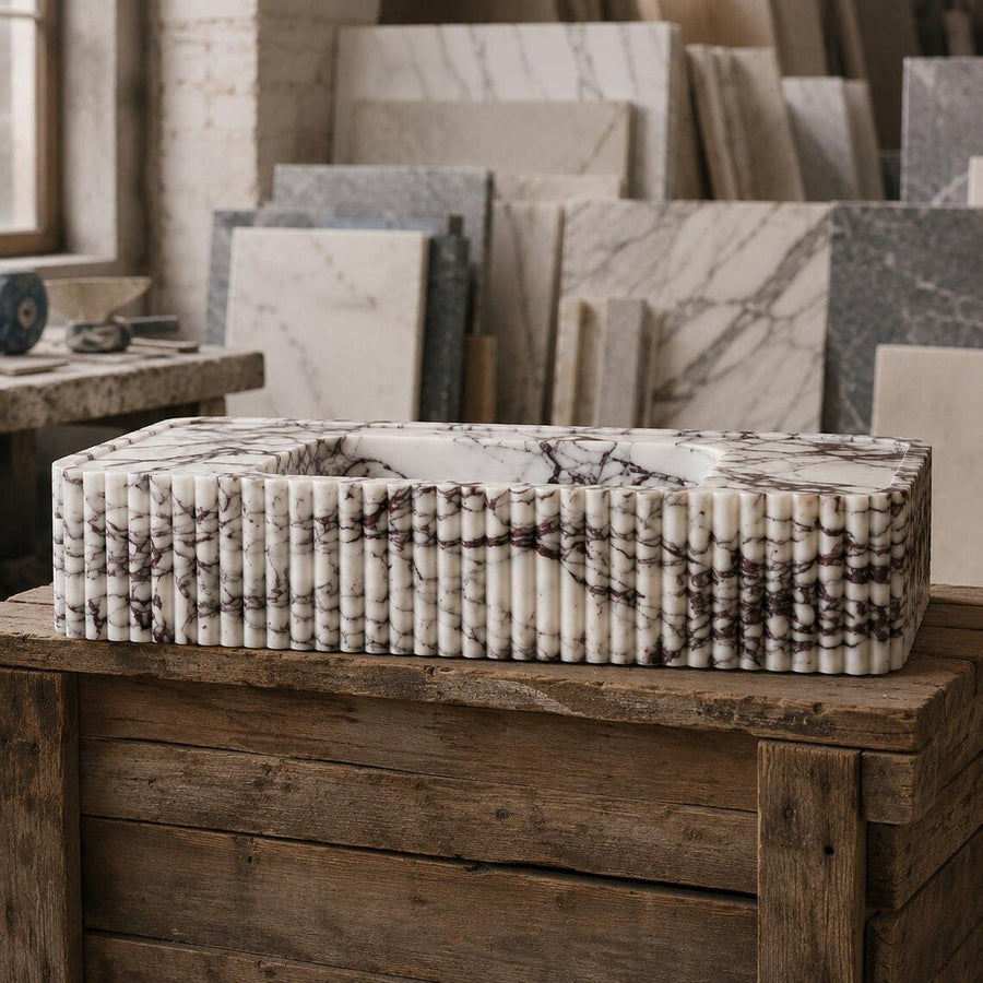A rectangular marble sink vessel with a ribbed, fluted exterior and a central basin, displayed on a rustic wooden crate in a stone workshop setting.