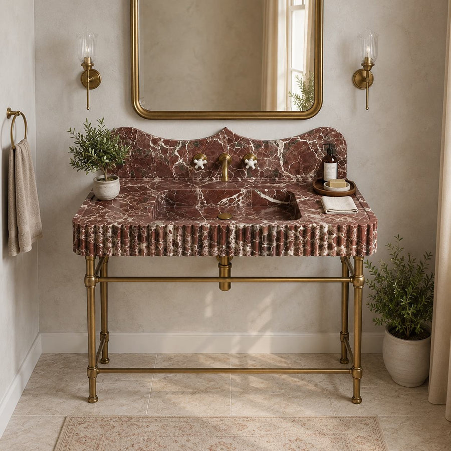 A luxury bathroom vanity featuring a rust-red and white veined marble countertop and sink basin with a scalloped backplate, supported by an ornate brass stand. The vanity is styled in a warm, elegant bathroom with a gold-framed mirror, brass sconces, a hanging towel, and potted plants.