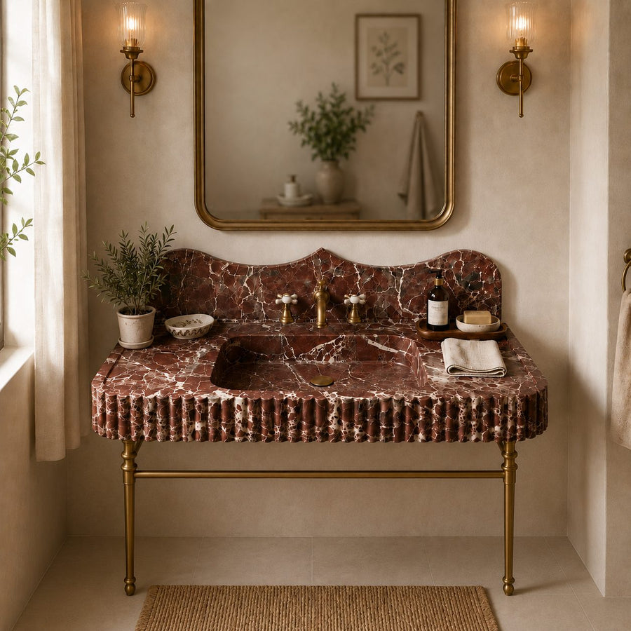 A luxury bathroom vanity featuring a dark reddish-brown marble sink and matching marble backsplash with gold brass legs and fixtures, set in a warm beige room with a gold-framed mirror and wall sconces.