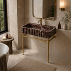 A modern bathroom vanity setup featuring a rectangular dark brown marble sink with a ribbed edge and a polished gold metal stand, faucet, and mirror, set in a warm beige stone bathroom.