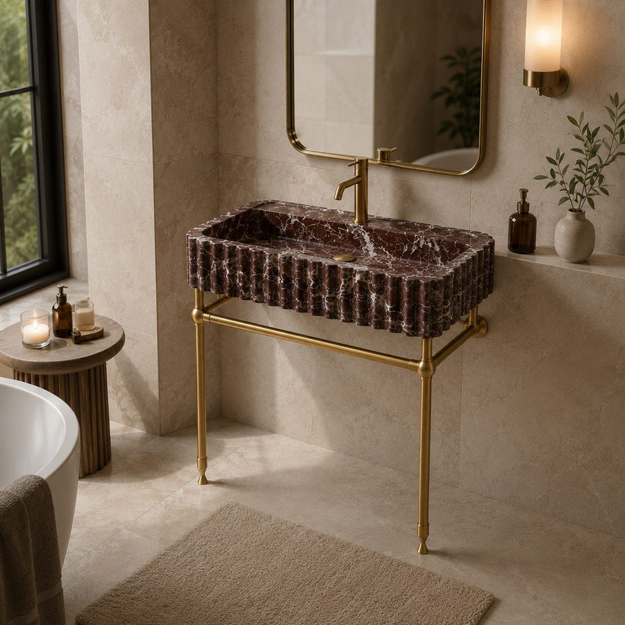 A modern bathroom vanity setup featuring a rectangular dark brown marble sink with a ribbed edge and a polished gold metal stand, faucet, and mirror, set in a warm beige stone bathroom.