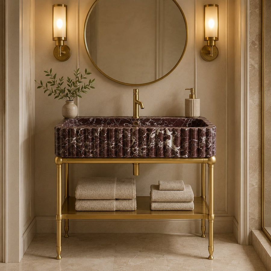 A luxury bathroom vanity featuring a rectangular ribbed purple marble sink with gold hardware, set on a gold metal console table with folded towels below. The background includes a round gold-framed mirror, two glowing wall sconces, and a small plant.