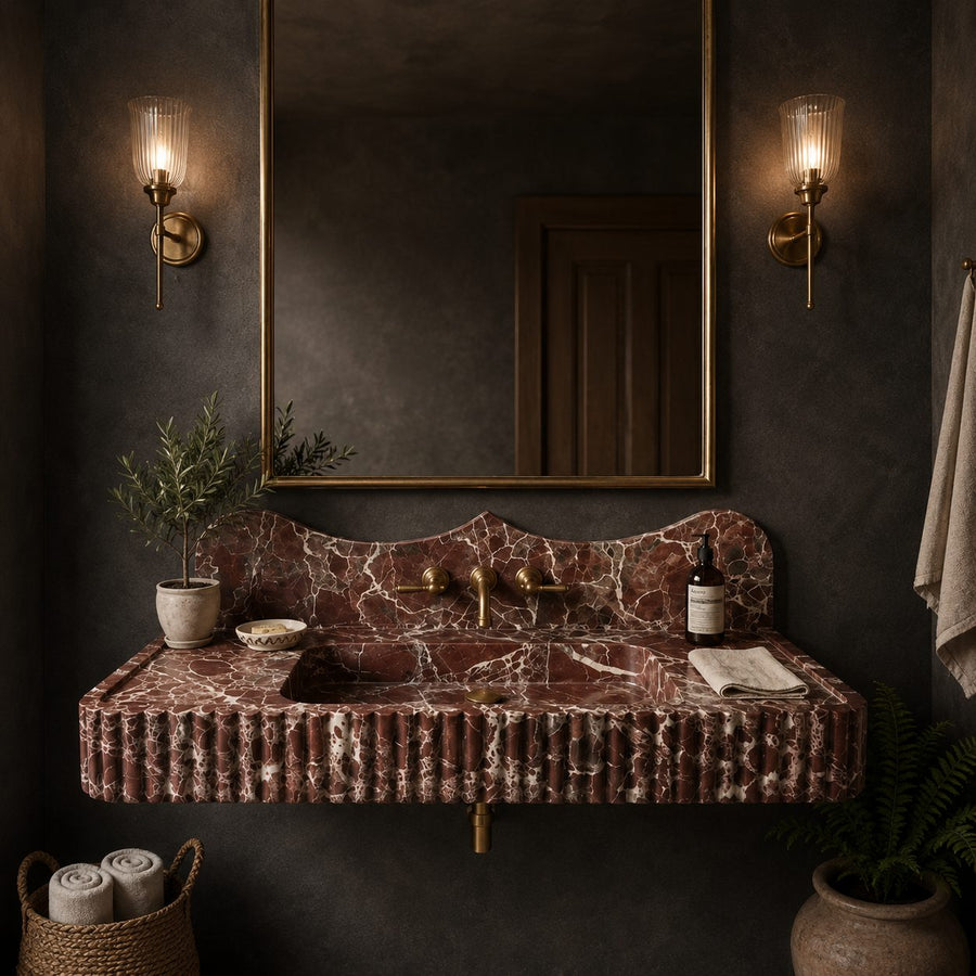 A wall-mounted sink vanity in a dark bathroom setting, featuring a ribbed terracotta-red marble countertop and matching marble backsplash, with a large gold-framed mirror and two lit wall sconces.