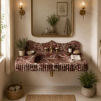 A floating bathroom vanity made of reddish-brown marble with a matching backsplash, set in a warm beige bathroom with brass fixtures, a gold-framed mirror, and potted plants.