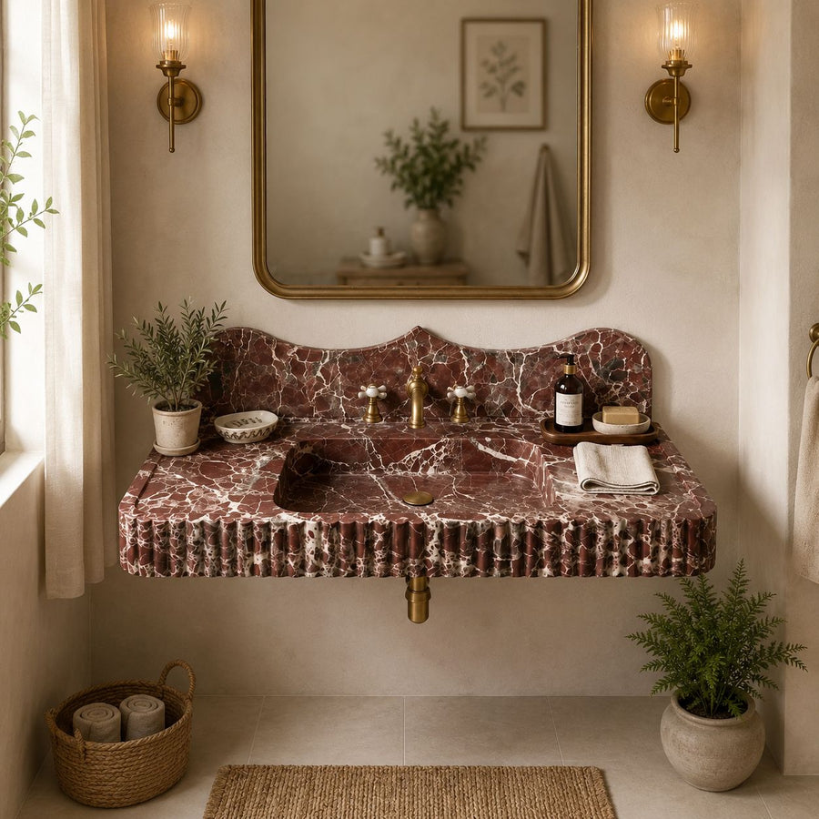 A floating bathroom vanity made of reddish-brown marble with a matching backsplash, set in a warm beige bathroom with brass fixtures, a gold-framed mirror, and potted plants.