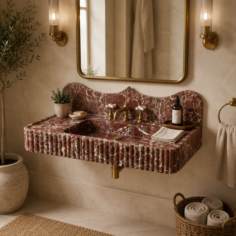 A wall-mounted bathroom sink with a sculptural fluted basin and a matching scalloped marble backsplash in reddish-brown and cream veining, installed in a warm beige bathroom with a gold-framed mirror, wall sconces, and toiletries.