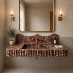 A floating bathroom vanity made of pinkish-brown marble with white veining, featuring a fluted front, integrated sink, and brass fixtures. The vanity is mounted against a textured wall beneath a large gold-framed mirror, flanked by two lit brass sconces.