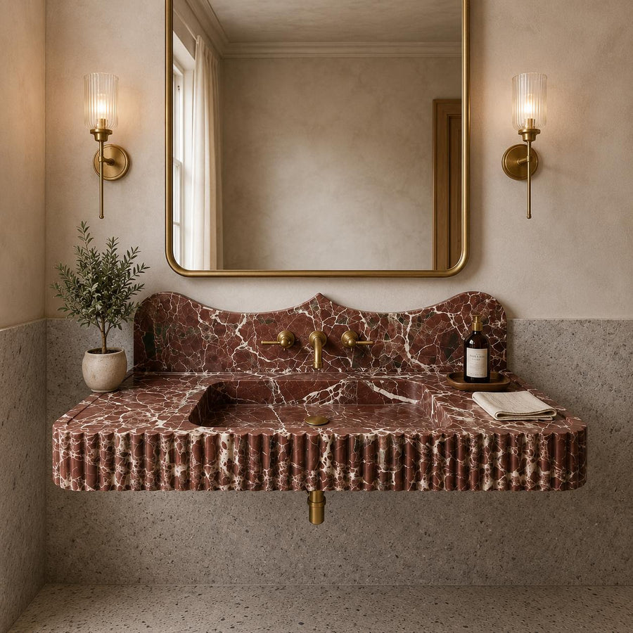 A floating bathroom vanity made of pinkish-brown marble with white veining, featuring a fluted front, integrated sink, and brass fixtures. The vanity is mounted against a textured wall beneath a large gold-framed mirror, flanked by two lit brass sconces.