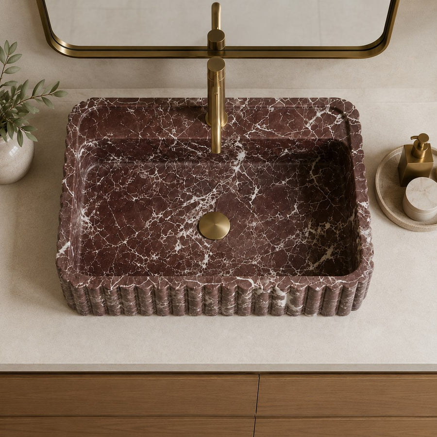 A rectangular vessel sink made of dark reddish-brown marble with white veining, featuring a fluted edge and a central drain. It is shown installed on a light beige countertop with a matching gold-finished faucet, a mirror, a small potted plant, and a soap dispenser.