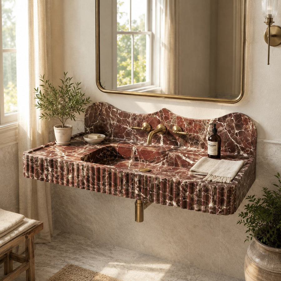 A floating bathroom sink made of reddish-brown marble with white veining, featuring an integrated sink, gold-tone faucet, mirror, and small decorative plants in a softly lit bathroom.