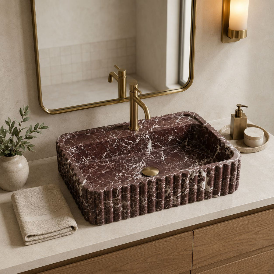 A rectangular vessel sink made of dark reddish-brown marble with white veining, set into a light stone countertop with a gold faucet and brass-toned hardware. A folded beige towel, a small potted plant, and a soap dispenser are visible on the counter, with a wall mirror and warm wall sconce in the background.