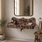 A wall-mounted bathroom sink with a scalloped edge and ribbed apron front, made of reddish-brown marble with white veining. It is shown installed on a beige plaster wall beneath a gold-framed mirror, with brass fixtures, a small wooden stool, a towel ring, and a potted plant in a styled bathroom setting.