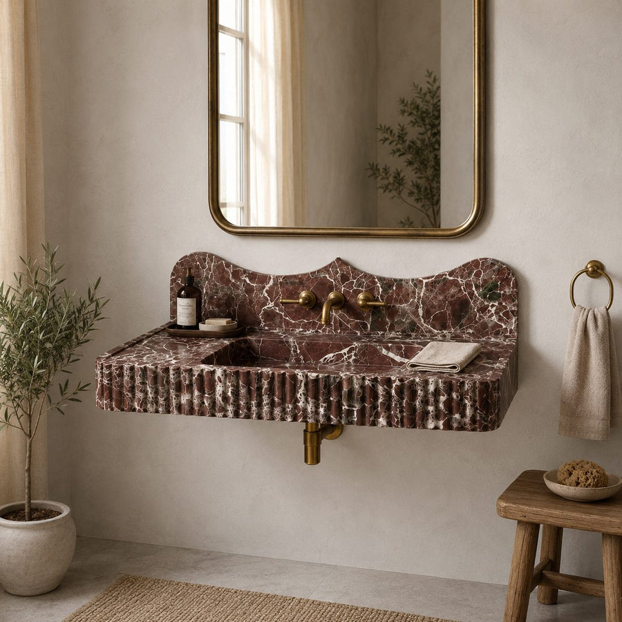 A wall-mounted bathroom sink with a scalloped edge and ribbed apron front, made of reddish-brown marble with white veining. It is shown installed on a beige plaster wall beneath a gold-framed mirror, with brass fixtures, a small wooden stool, a towel ring, and a potted plant in a styled bathroom setting.