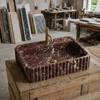 A rectangular dark red marble vessel sink with fluted edges and a brass faucet, displayed on a rustic wooden workbench in a stone and tile workshop.