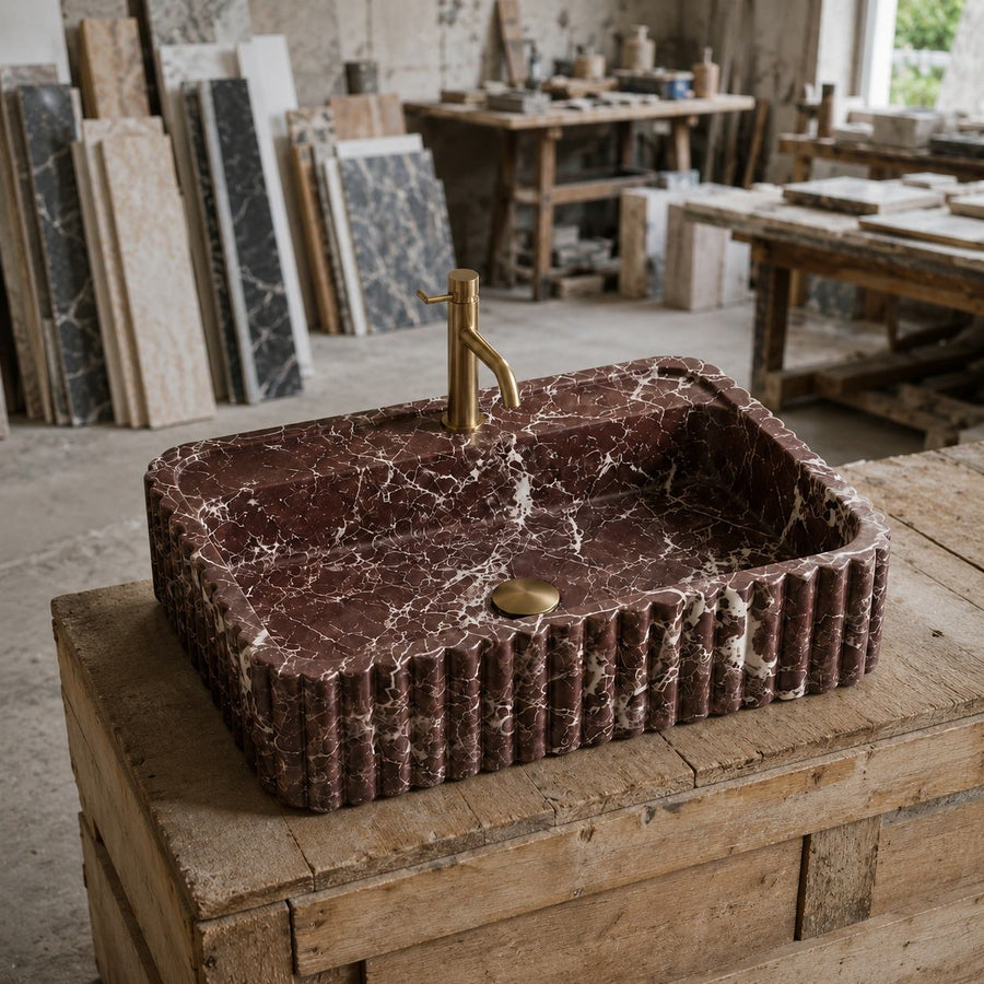 A rectangular dark red marble vessel sink with fluted edges and a brass faucet, displayed on a rustic wooden workbench in a stone and tile workshop.
