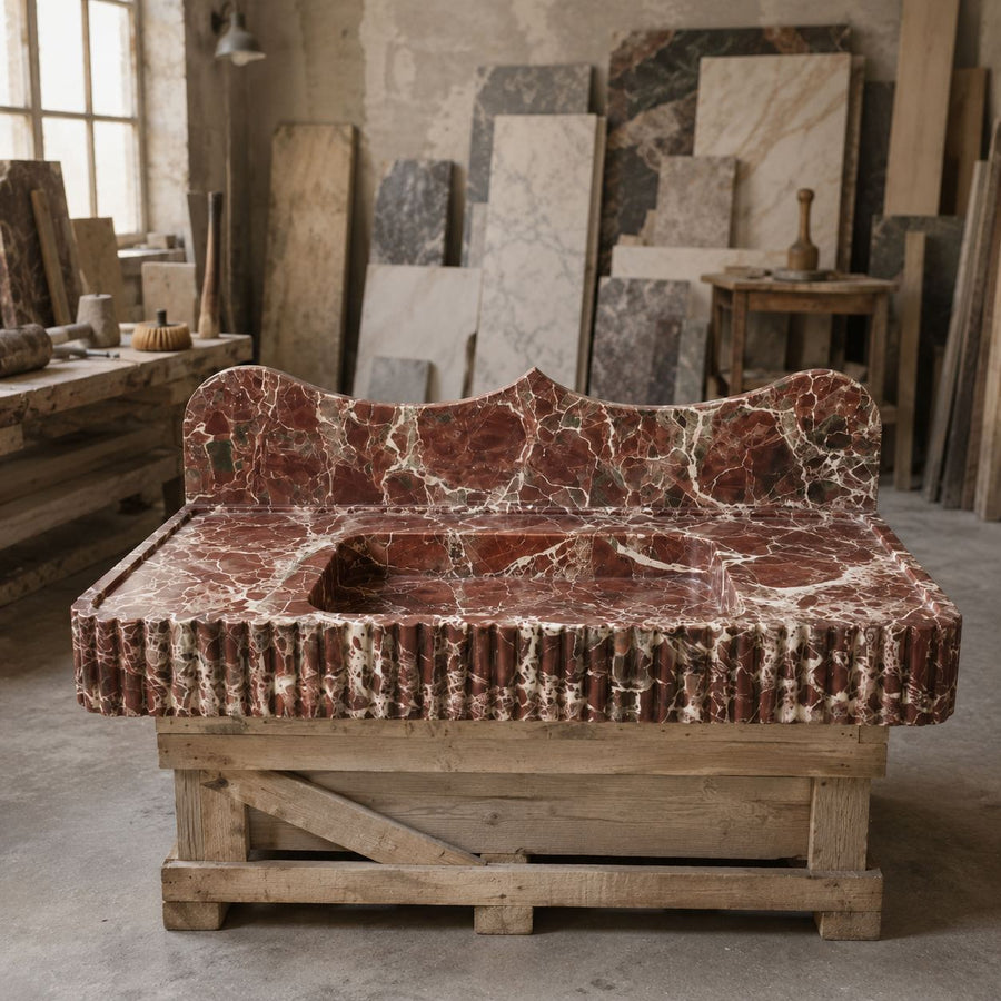 A carved stone bench or daybed made from reddish-brown marble with white veining, displayed on a rustic wooden support frame inside a stone workshop.