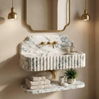 A floating bathroom vanity made of white and gray marble with fluted edges, mounted on a beige wall beneath a gold-framed mirror. The vanity includes a matching marble shelf with folded towels and small decor items, and gold-tone faucet and fixture details.
