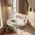 A floating marble sink with fluted edges and a curved backsplash, paired with a gold faucet and gold-framed wall mirror, shown in a styled bathroom setting with warm sunlight and a small potted plant.