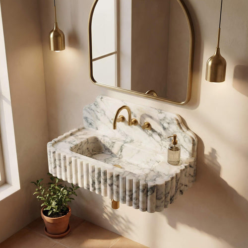 A floating marble sink with fluted edges and a curved backsplash, paired with a gold faucet and gold-framed wall mirror, shown in a styled bathroom setting with warm sunlight and a small potted plant.