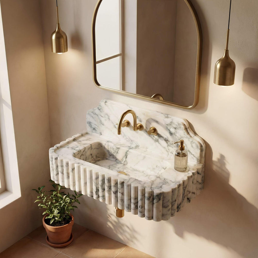 A floating marble sink with fluted edges and a curved backsplash, paired with a gold faucet and gold-framed wall mirror, shown in a styled bathroom setting with warm sunlight and a small potted plant.