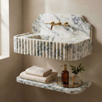 A wall-mounted marble sink with a fluted front and backplate, paired with a matching floating shelf holding folded towels, a brown pump bottle, and a small vase with greenery. The fixtures are brushed gold, set against a warm beige wall.
