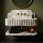 A modern double-sink bathroom vanity made of white marble with gray veining, featuring a fluted front, a shelf below, and brass fixtures. The vanity is styled with black towels, black soap dispensers, and a lit candle against a dark olive green wall.