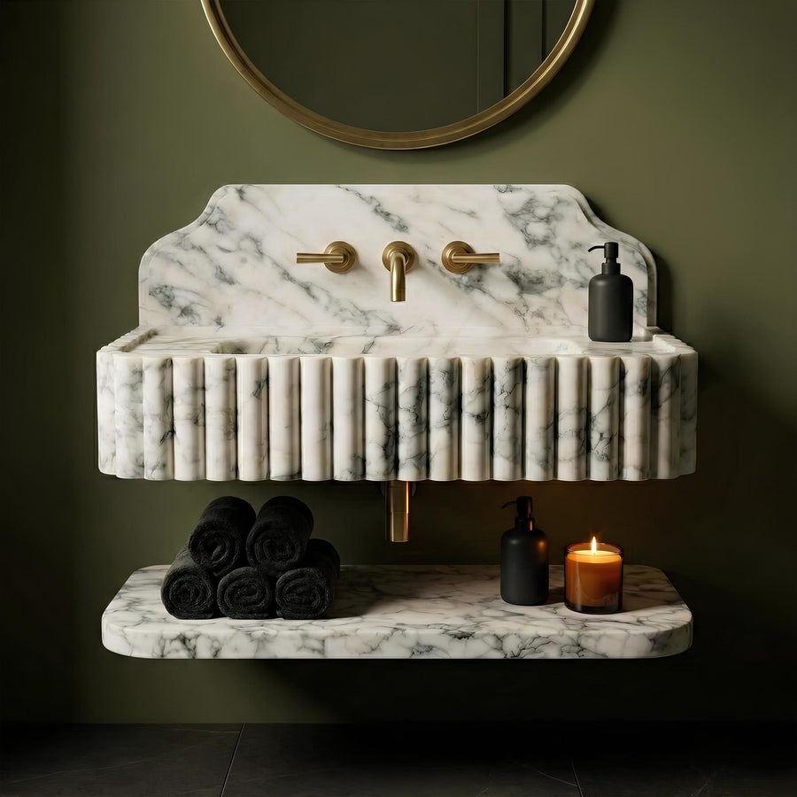 A modern double-sink bathroom vanity made of white marble with gray veining, featuring a fluted front, a shelf below, and brass fixtures. The vanity is styled with black towels, black soap dispensers, and a lit candle against a dark olive green wall.