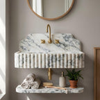 A wall-mounted bathroom sink made of white marble with gray veining, featuring a fluted front, a curved backplate, a gold faucet, and a matching marble shelf below holding rolled towels, a soap dispenser, a brush, and a small potted plant.