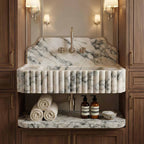 A luxury bathroom vanity featuring a fluted white marble sink basin with gray veining, a matching marble backsplash, and a fluted front apron. The vanity is flanked by dark wood cabinets with brass handles, and the open shelf below holds three rolled white towels, two amber pump bottles, and a small bowl.