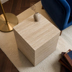 Beige stone side table with a vase on a beige rug in a room with a blue chair and gold lamp.