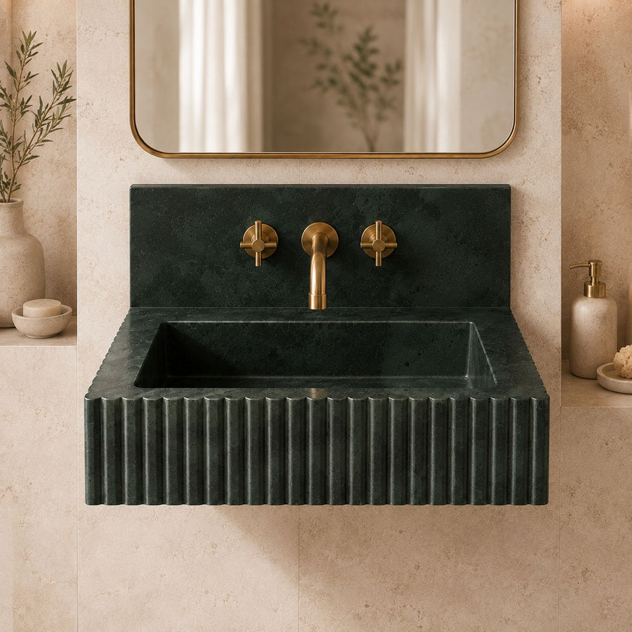A dark green fluted stone sink with a matching backplate and brass faucet, shown in a styled bathroom setting with a mirror and beige wall.