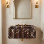 A wall-mounted rectangular bathroom sink with a dark purple marble fluted exterior and gold faucet hardware, shown below a matching gold-framed mirror with two gold sconces on a beige wall.