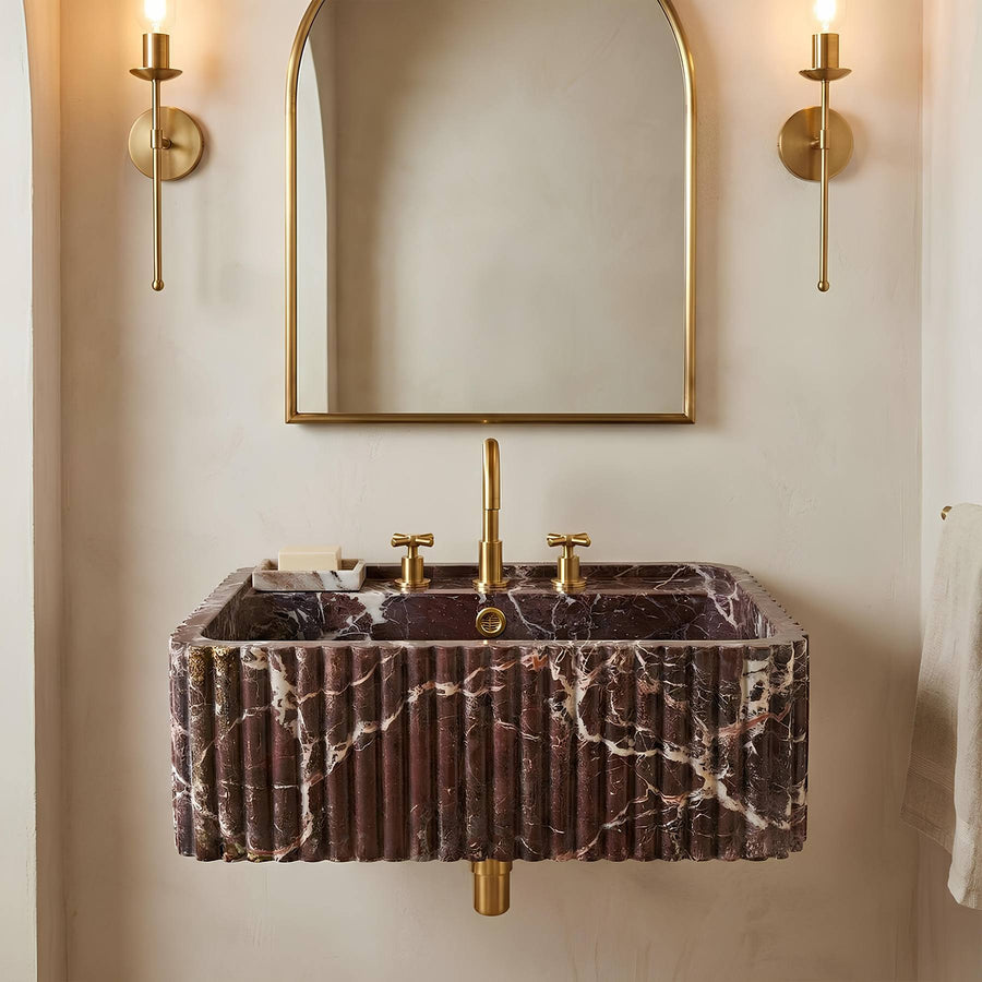 A wall-mounted rectangular bathroom sink with a dark purple marble fluted exterior and gold faucet hardware, shown below a matching gold-framed mirror with two gold sconces on a beige wall.