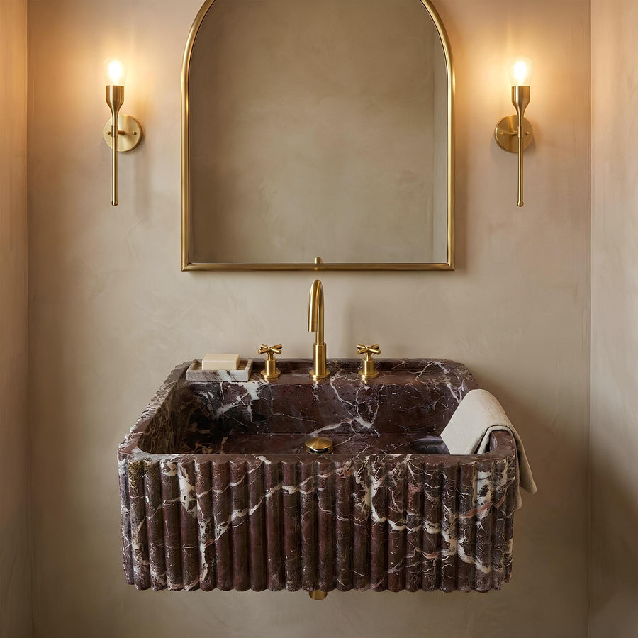 A luxury wall-mounted bathroom sink made of dark brown marble with white veining, featuring a fluted front, a tall gold faucet, and gold fixtures. It is styled in a bathroom with a matching gold-framed arched mirror and two gold sconces on a beige plaster wall.